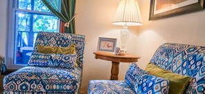 Una habitación con sillas estampadas, una pequeña mesa de madera con una lámpara y un cuadro enmarcado.