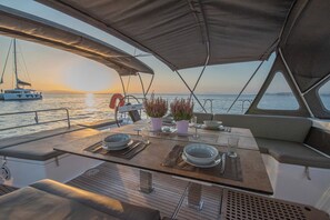 A dining area on a boat with a table set for two, a potted plant, and a view of the sunset over the water. - Sun-Kissed Sailing Adventure in Islas Sarónicas (Alimos)