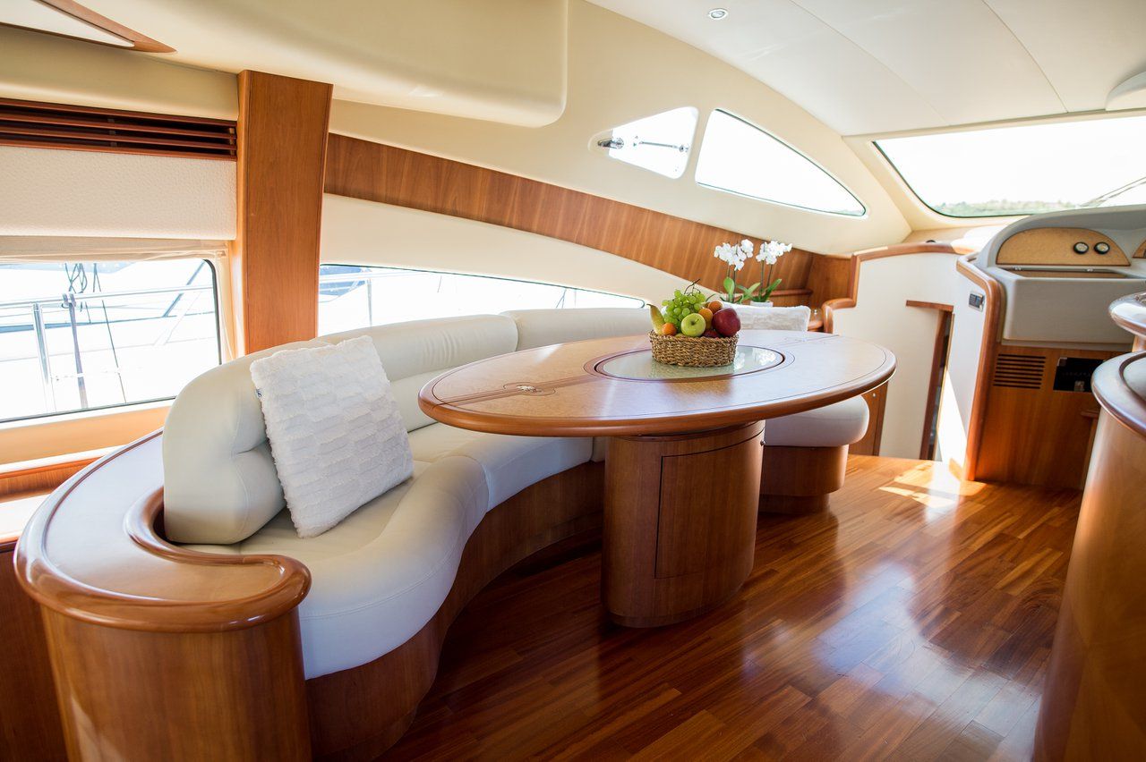 A yacht interior with a curved white sofa, a round wooden table with a fruit basket, and a small kitchen area.