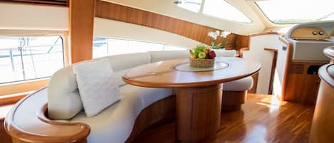 Interieur einer Yacht mit einer gebogenen weißen Couch, einem runden Holztisch mit Obstschale und einer kleinen Küchenzeile.