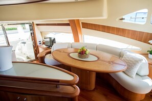 A boat interior with a round table, cushioned seating, a small plant, and a window.