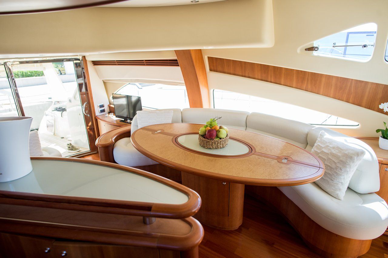 A boat interior with a round table, cushioned seating, a small plant, and a window.