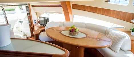 A boat interior with a round table, cushioned seating, a small plant, and a window.
