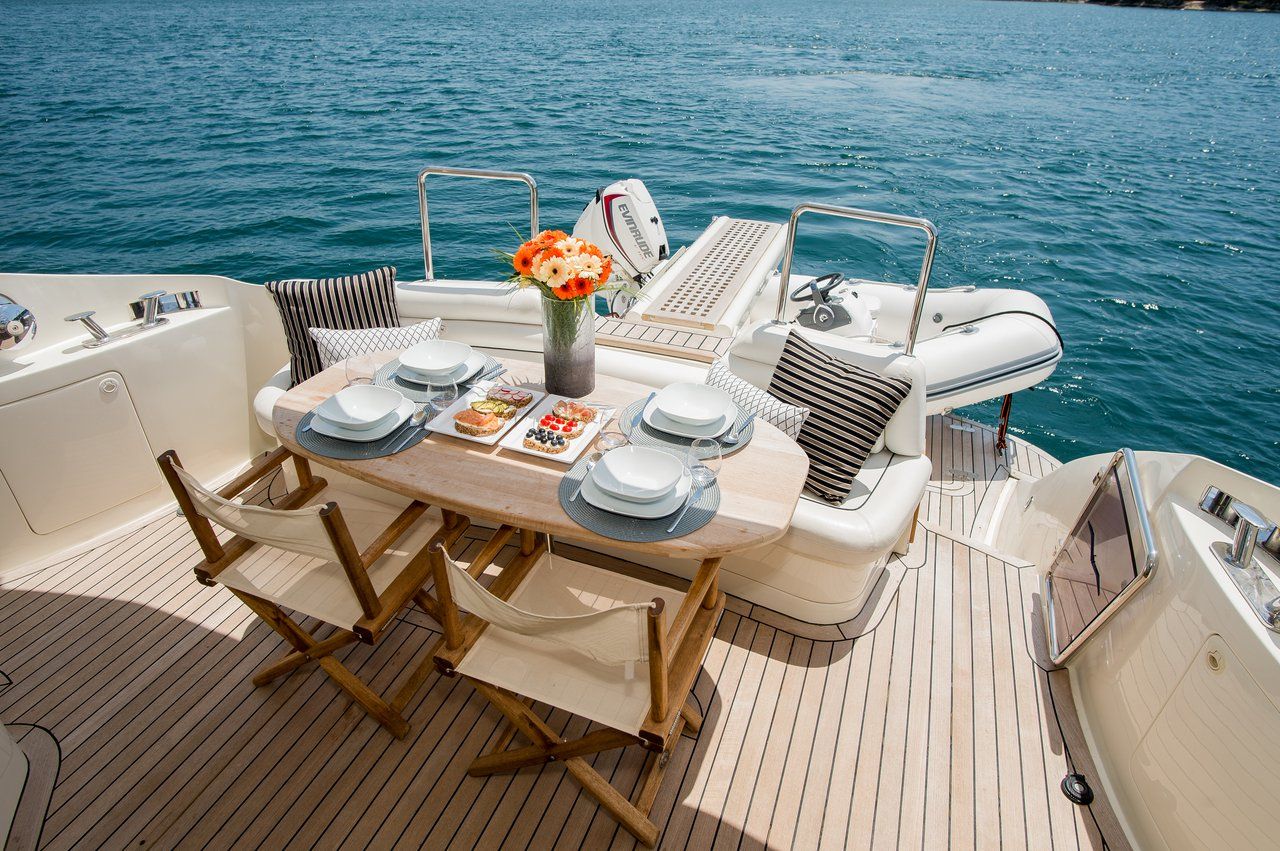 A boat deck with a dining table set for four, surrounded by cushioned chairs, a small table with a vase of flowers, and a grill.