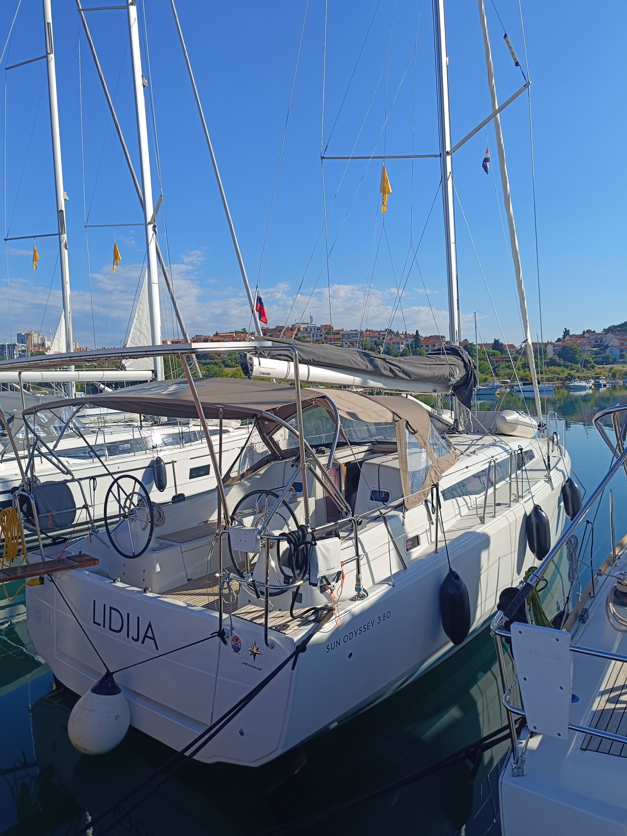 Ein Segelboot liegt in einer Marina vor blauem Himmel, im Hintergrund sind Gebäude zu sehen.