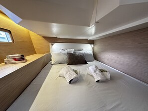 A compact hotel room with a bed, pillows, a bedside table with books, and a window. - Sun-Kissed Sailing Adventure in Islas Sarónicas (Alimos)