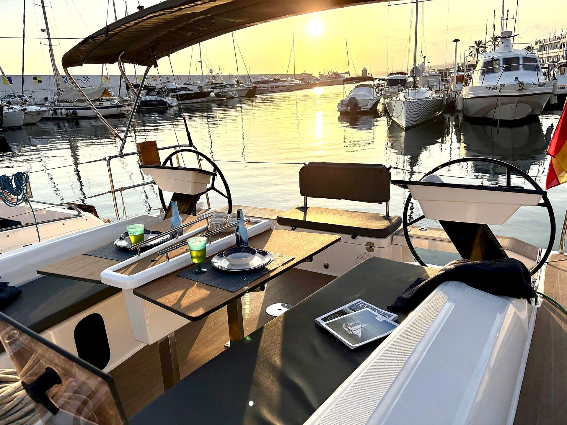 Un port de plaisance avec des bateaux amarrés, une table avec un ordinateur portable, et un coucher de soleil en arrière-plan.