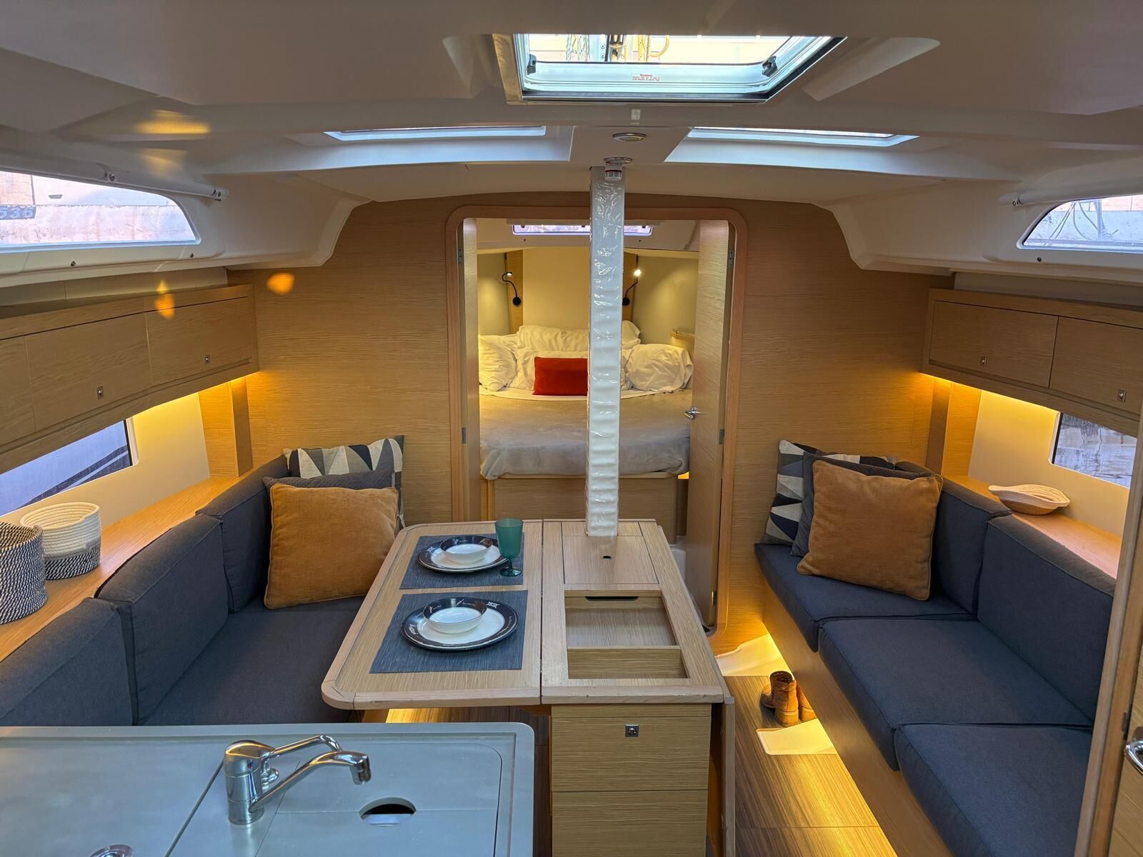 Een compact interieur met een centrale eettafel, twee banken, een bed en dakramen.