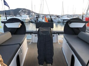 A boat deck with two cushioned seating areas, a flag, and a view of a marina with boats and buildings.