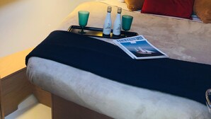 Una stanza compatta con un letto, un tavolino con bottiglie e bicchieri, una rivista e un lucernario.