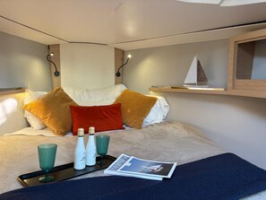 A neatly made bed with pillows, a tray with bottles and glasses, a magazine, and a small sailboat model on a shelf.