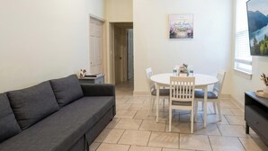 A living room with a grey sofa, a dining table with chairs, a TV on the wall, and a door leading to another room.