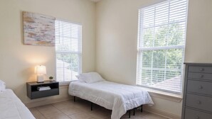A bedroom with two beds, a nightstand with a lamp, a dresser, and large windows with blinds.