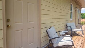 A wooden deck with two chairs and a small table, a beige door with a brass doorknob, and a window with a white frame.