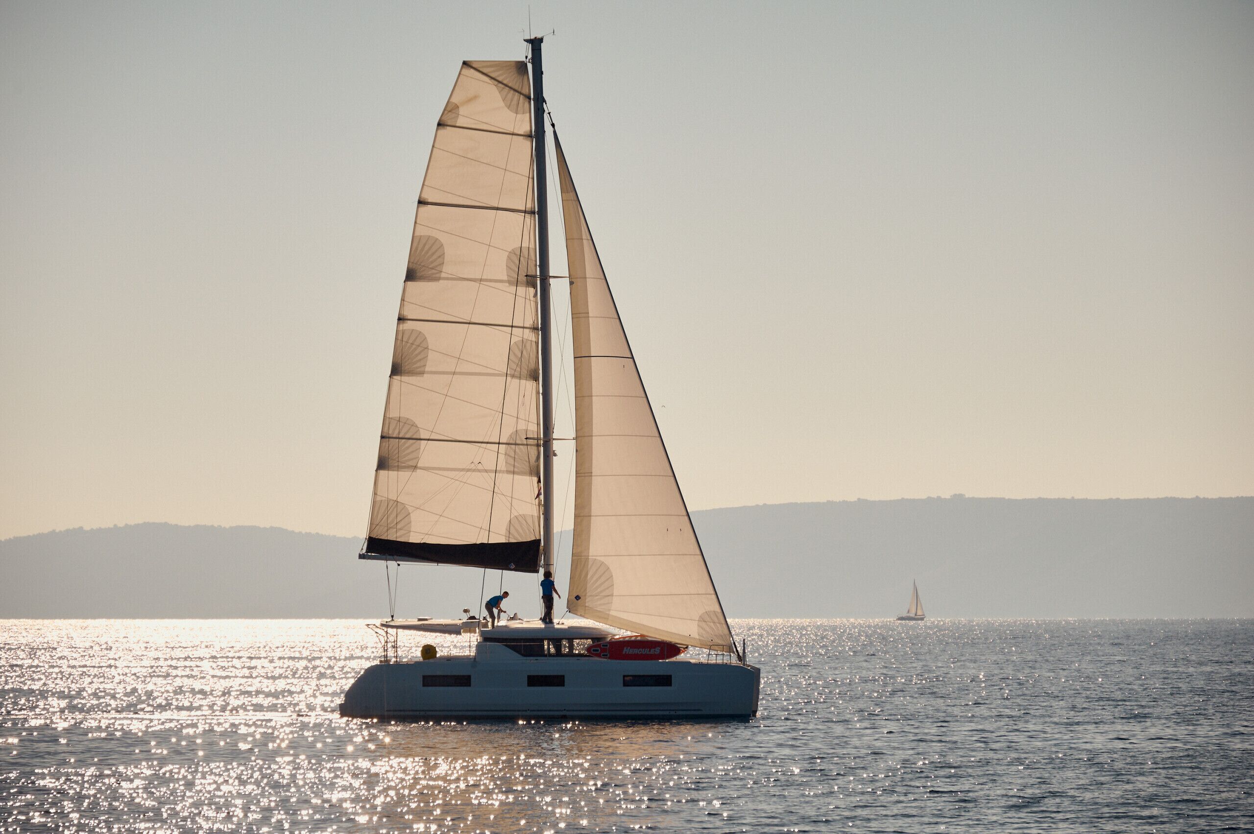 Un velero con una persona de pie en la cubierta, otra persona cerca de la popa y un velero distante en el horizonte.