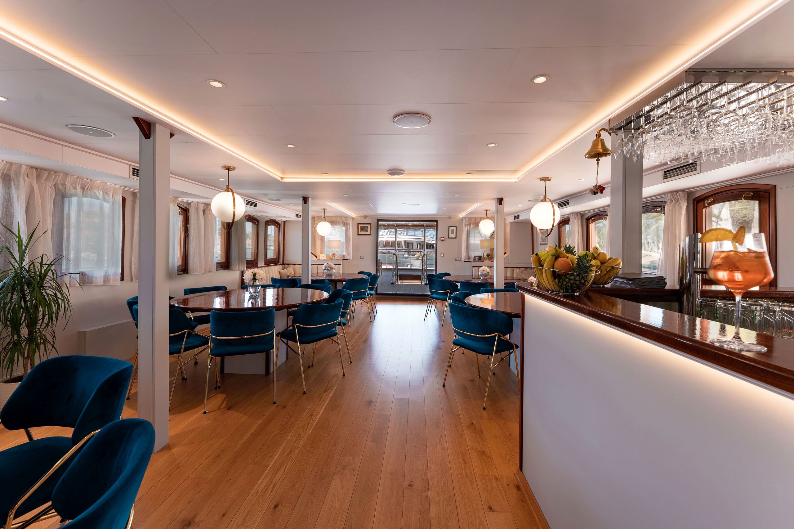 A hotel dining area with round tables, blue upholstered chairs, and a bar counter with a fruit bowl and glasses.