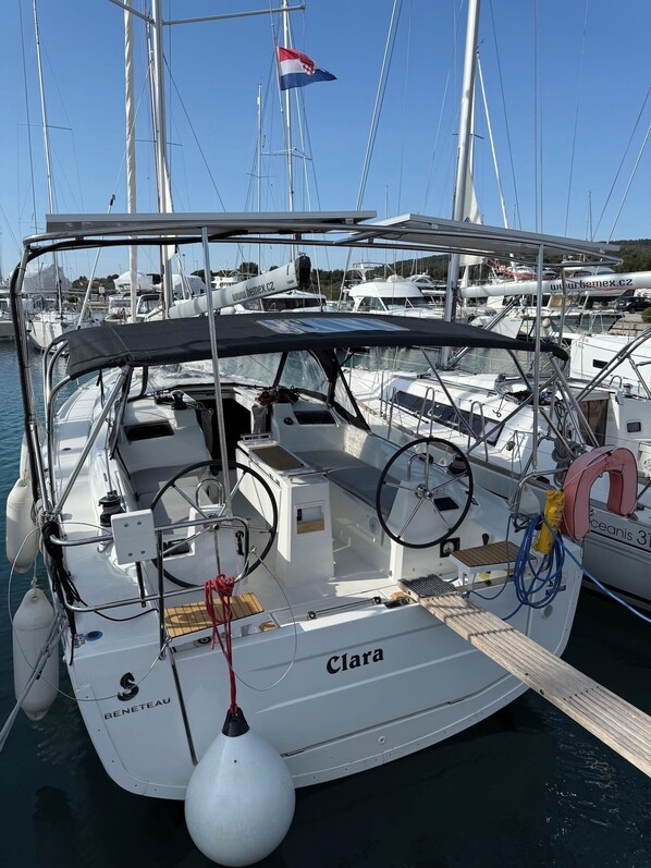 Un velero llamado 'Clara' atracado en un puerto deportivo junto a otros barcos, una bandera y un banner del sitio web.