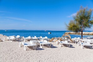 Ein Strand mit weißen Kunststoffliegen, die in Reihen angeordnet sind, eine Palme und eine felsige Küstenlinie im Hintergrund mit Booten.