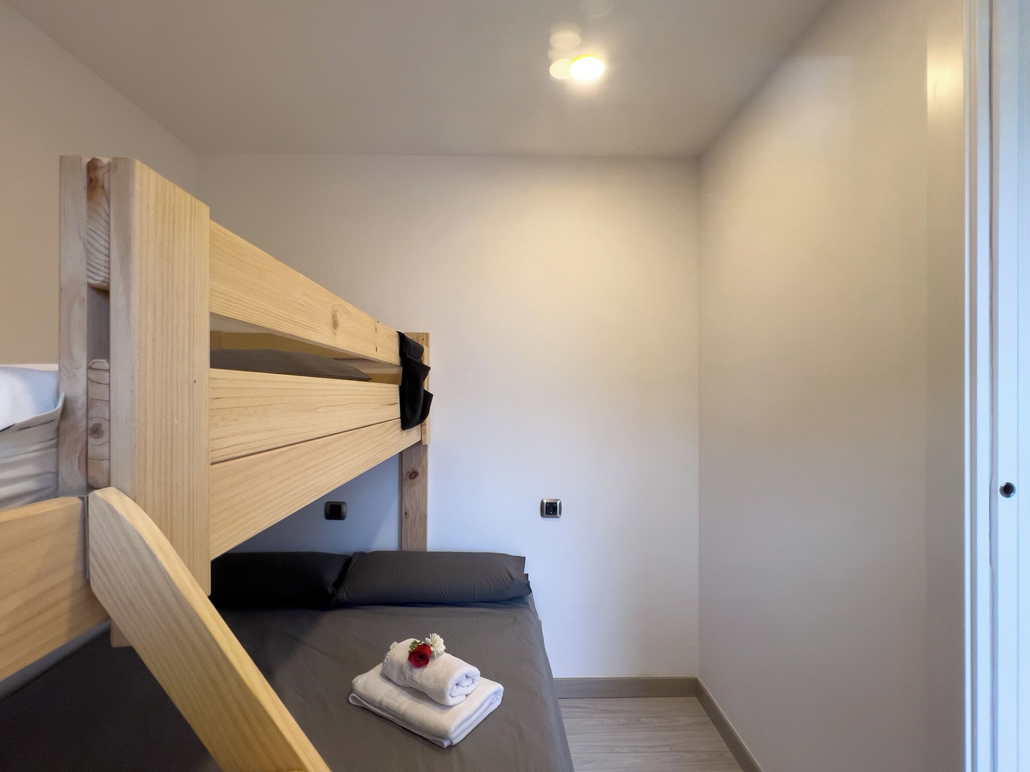 A bunk bed with a small bedside table, a single bed with a pillow, and a neatly folded towel with a flower on the bed.