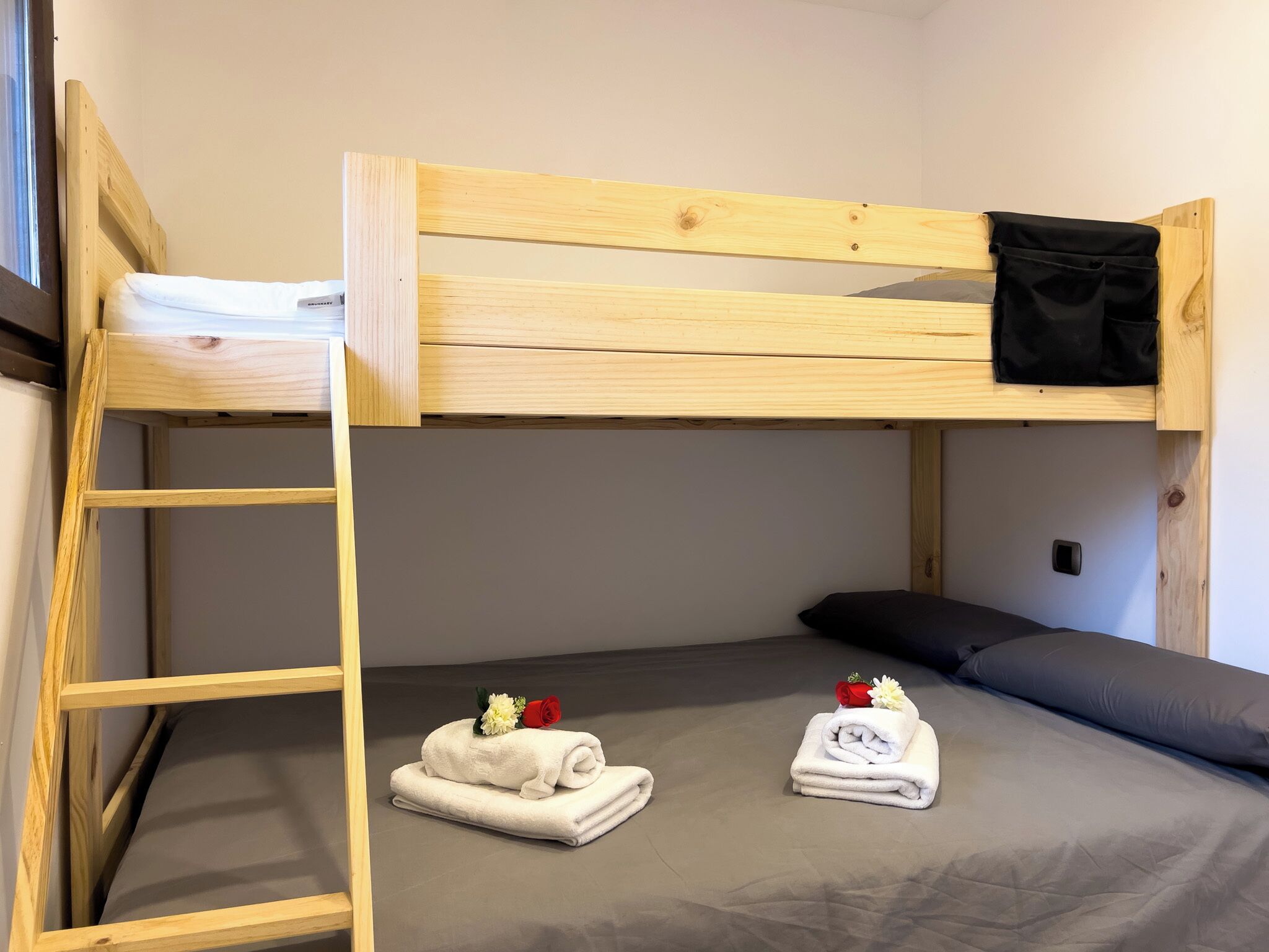 A bunk bed room with a wooden ladder, a gray mattress, two neatly folded white towels with flowers, and a black jacket on the top bunk.