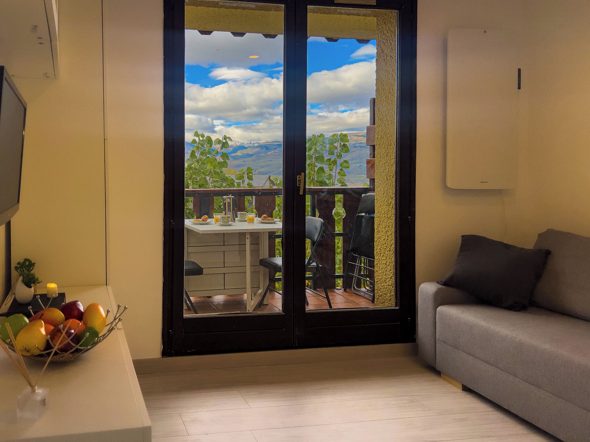 Una habitación con puerta corrediza de vidrio que da a un espacio exterior con una mesa y sillas, un sofá gris y un frutero a la izquierda.