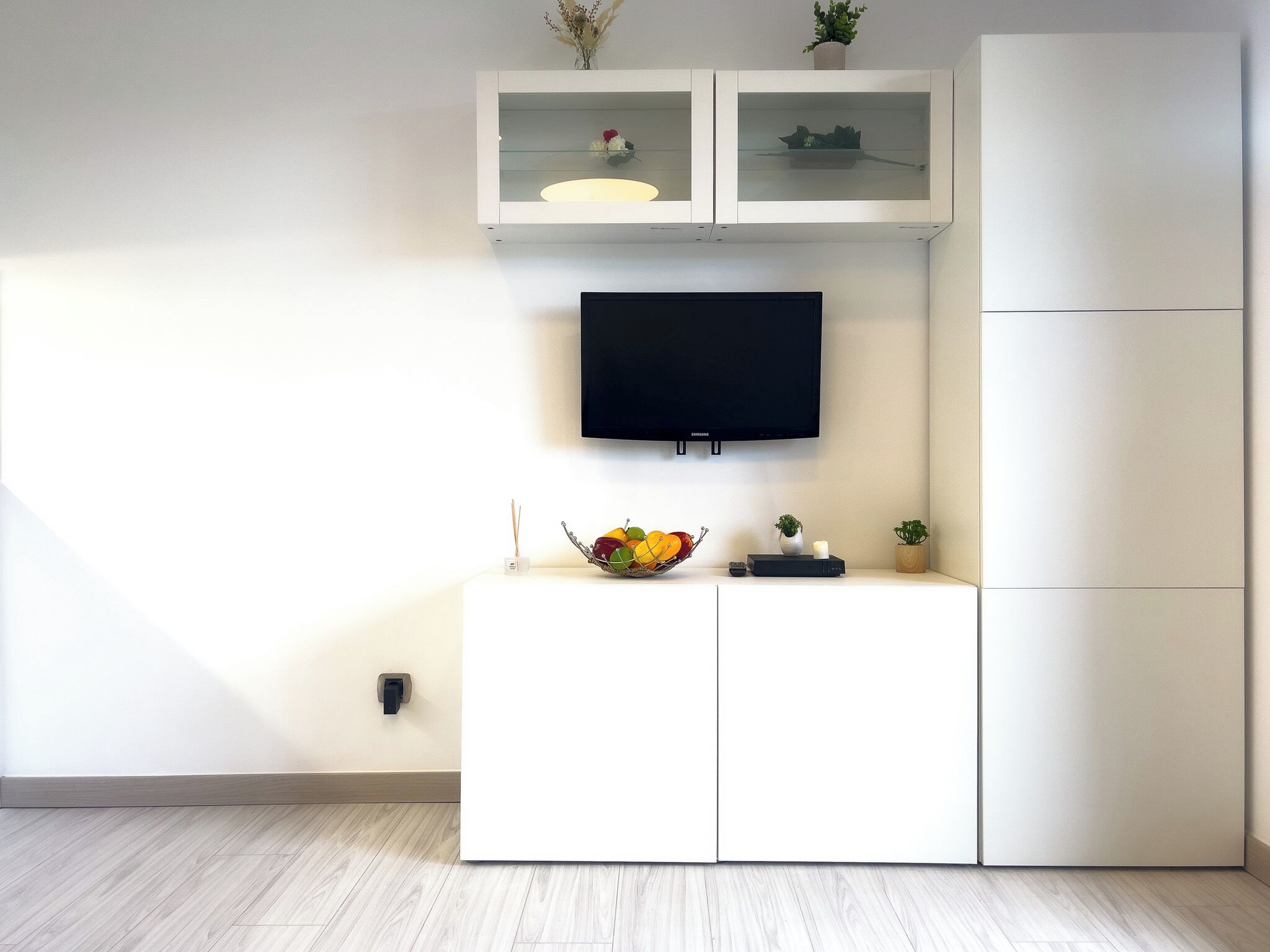 Un salon moderne avec un téléviseur à écran plat fixé au mur, un meuble blanc sur lequel est posé un panier de fruits, et une petite plante sur le dessus du meuble.