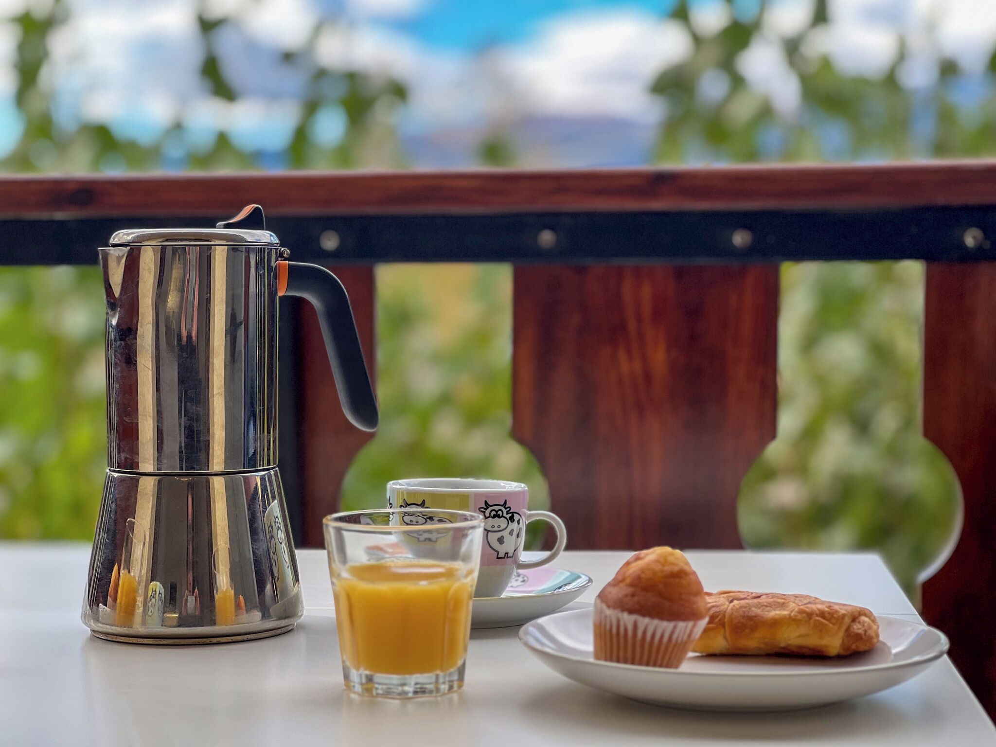 A table with a metallic coffee pot, a glass of orange juice, a mug, and a plate with a pastry.