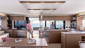 A modern kitchen with a large TV, a dining area with a table, and a view of the sea.