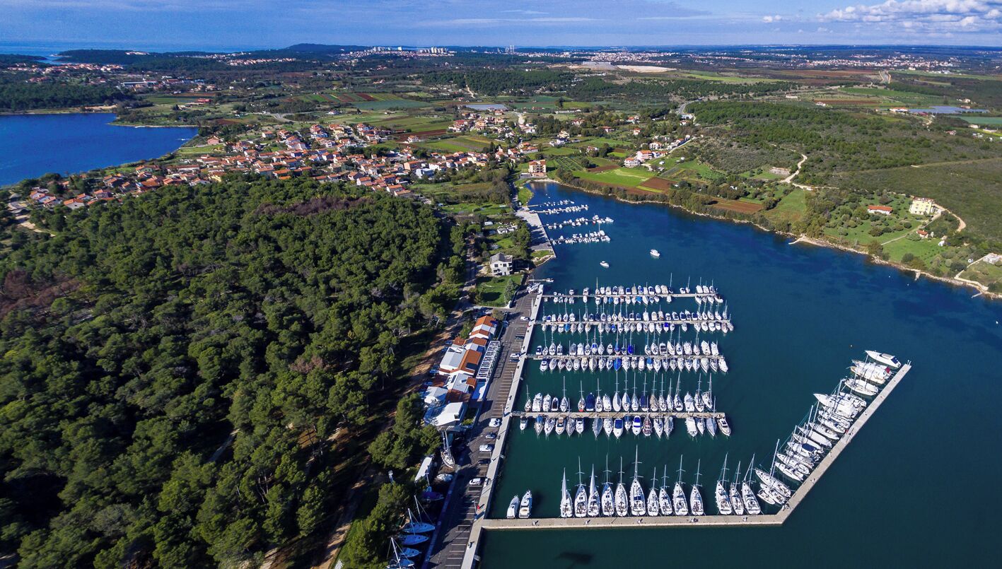Eine Vogelperspektive eines Yachthafens mit zahlreichen angestaubten Booten, umgeben von einem bewaldeten Gebiet und einer kleinen Stadt im Hintergrund.