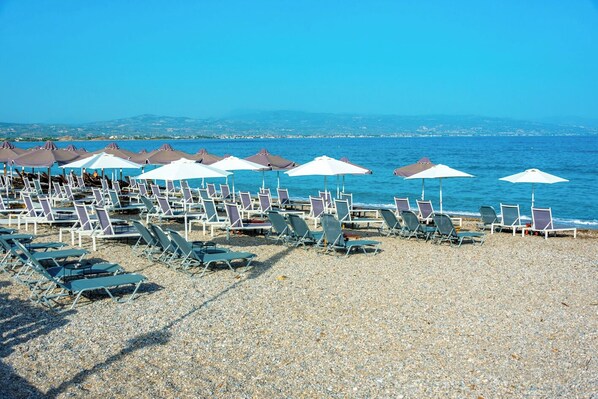 Une plage bordée de rangées de chaises longues et de parasols, une plage de galets, et la mer en arrière-plan.