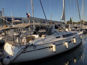 Eine weiße Yacht namens 'Cruiser 46' liegt in einer Marina festgemacht, zusammen mit anderen Booten, unter einem wolkenlosen Himmel, mit einem Gebäude im Hintergrund.