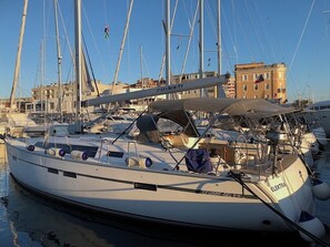 Un velero amarrado en un puerto deportivo con otros barcos, edificios al fondo y cielo despejado.