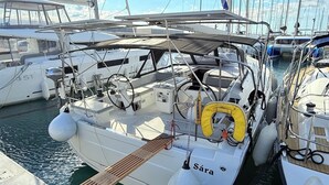 En segelbåt låg förtöjd vid en småbåtshamn, omgiven av flera andra båtar. På båten fanns ett ratt, en livboj och namnet ”Sára” var målat på skrovet.