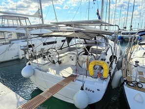 Eine Segelyacht liegt in einer Marina vor Anker, mit mehreren Booten, einem Steuerrad, einem Rettungsboje und dem Namen 'Sára' auf dem Rumpf.