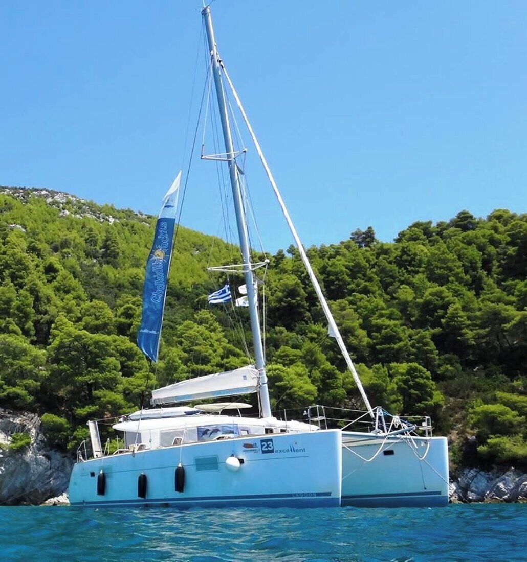 A sailboat with a flag, docked near a rocky coastline with dense green trees.