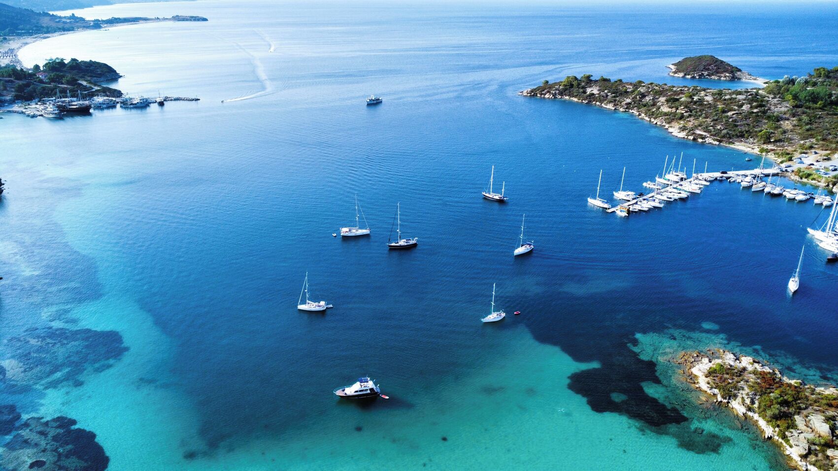 An aerial view of a coastal marina with multiple sailboats anchored in clear blue waters, surrounded by rocky terrain and a small island.