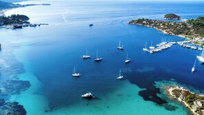 An aerial view of a coastal marina with multiple sailboats anchored in clear blue waters, surrounded by rocky terrain and a small island.