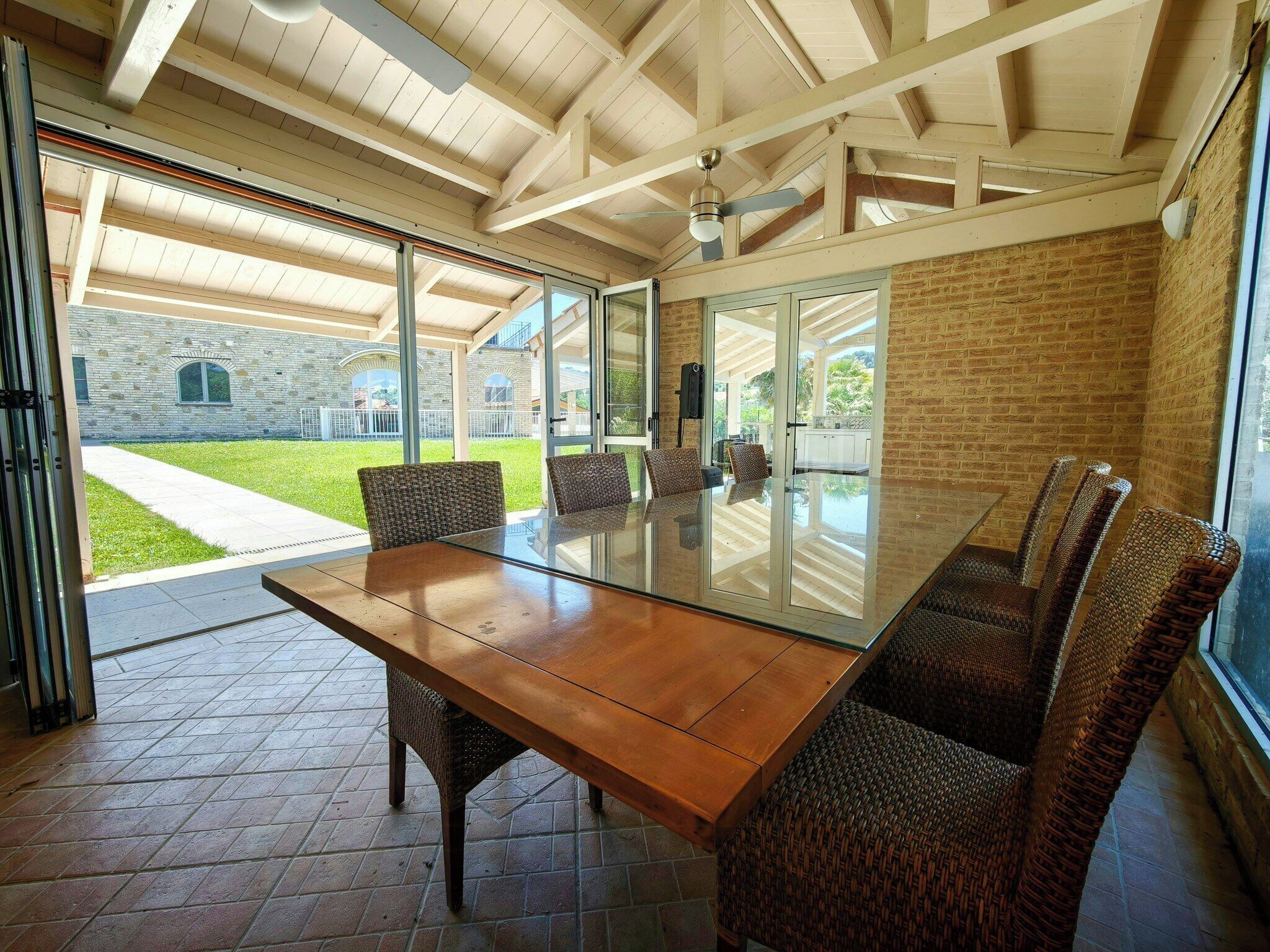 A spacious dining area with a glass table and wicker chairs, large windows, and a view of a brick building and lawn.