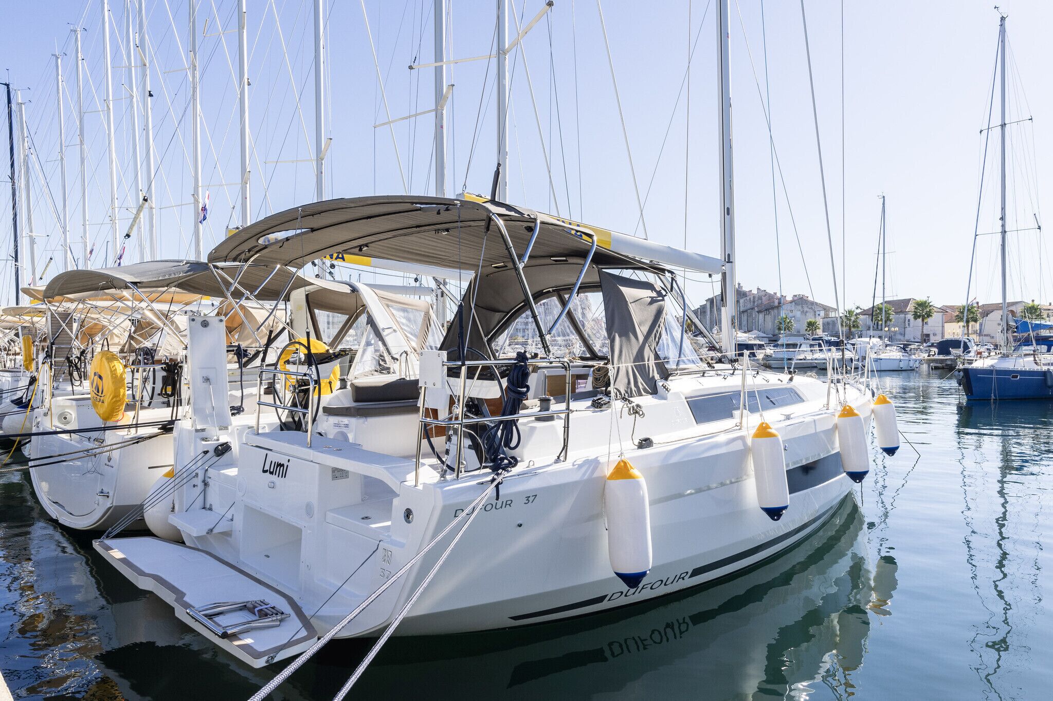 A white yacht docked at a marina with multiple masts, a covered deck, and a name 'Lumi' on the side.