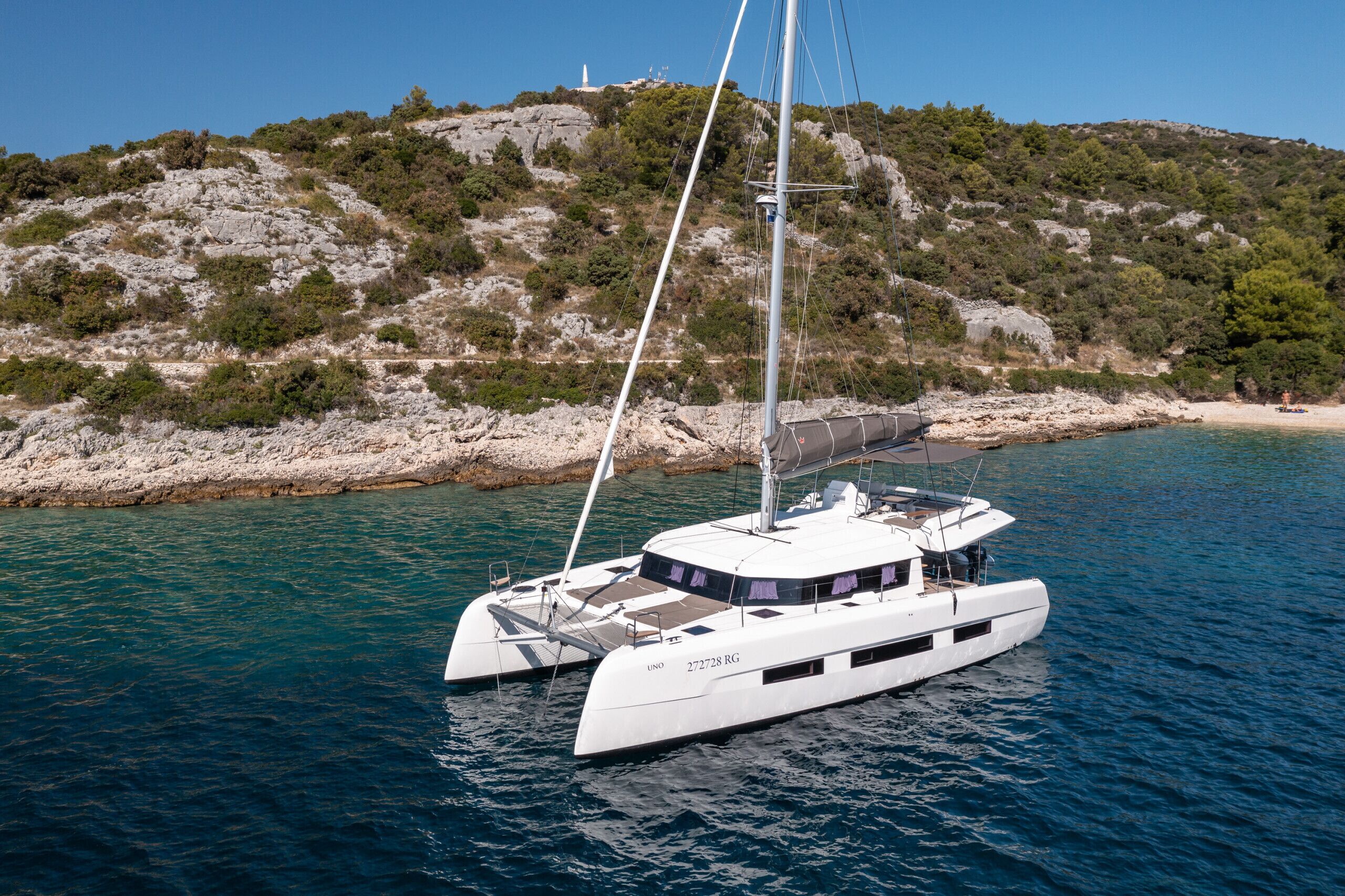 Un yacht blanc, doté d’une cabine et de voiles, est amarré près d’une côte rocheuse, avec un phare en arrière-plan.