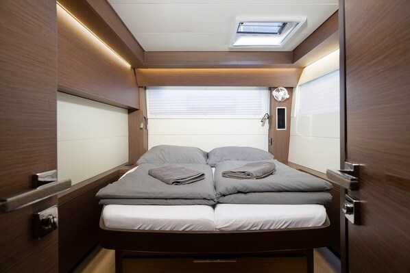 Interior compacto e bem iluminado, com uma cama, uma pequena janela e um ventilador.