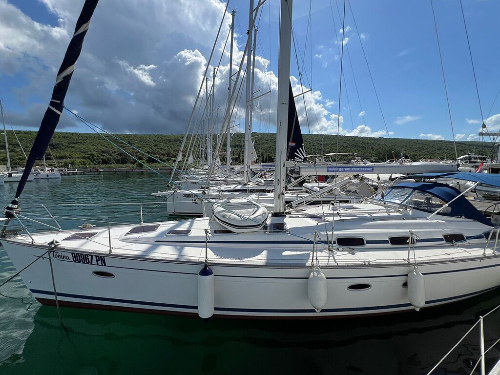 Un velero amarrado en una marina, junto a otros barcos, con un cielo despejado y un paisaje montañoso al fondo.