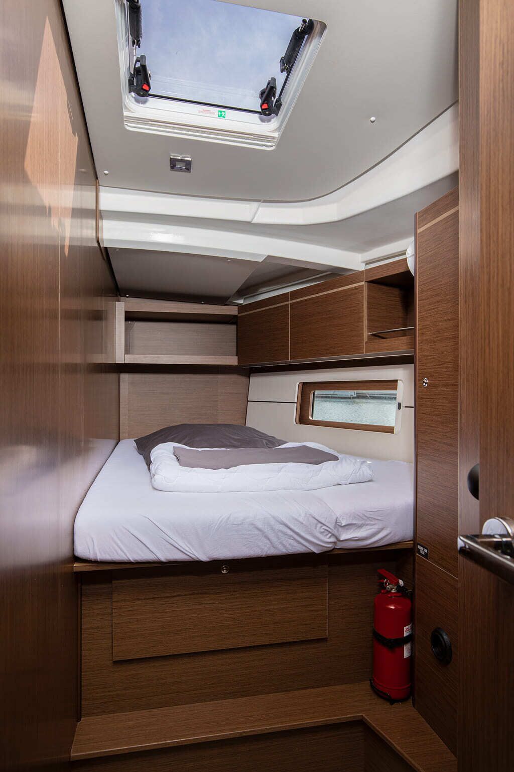 A compact, well-lit cabin with a skylight, a bed with pillows, a small window, and a fire extinguisher.