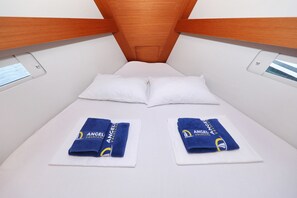 Cabine estreita e reservada, com teto de madeira, roupa de cama branca e duas toalhas com a marcação 'ANGEL Yacht'.