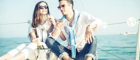 Two people on a boat, holding glasses, with a bottle and a metal container on the deck.