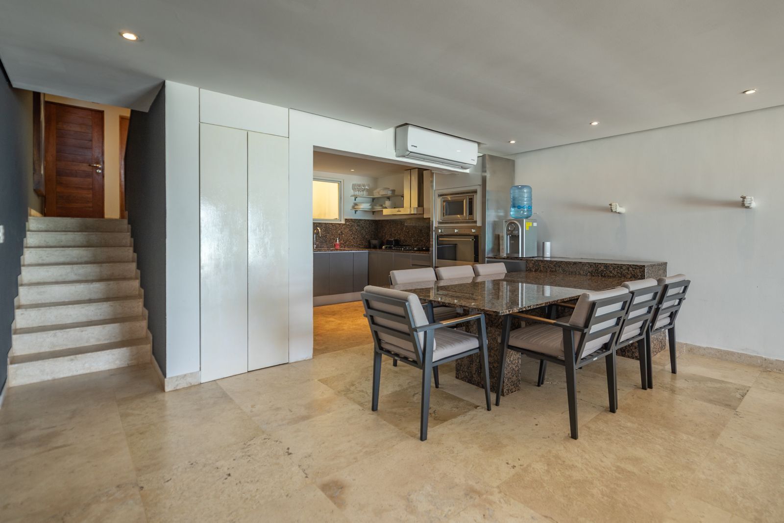 Una cocina moderna con una isla central, un área de comedor con una mesa grande y sillas, y una escalera que lleva a otro nivel.