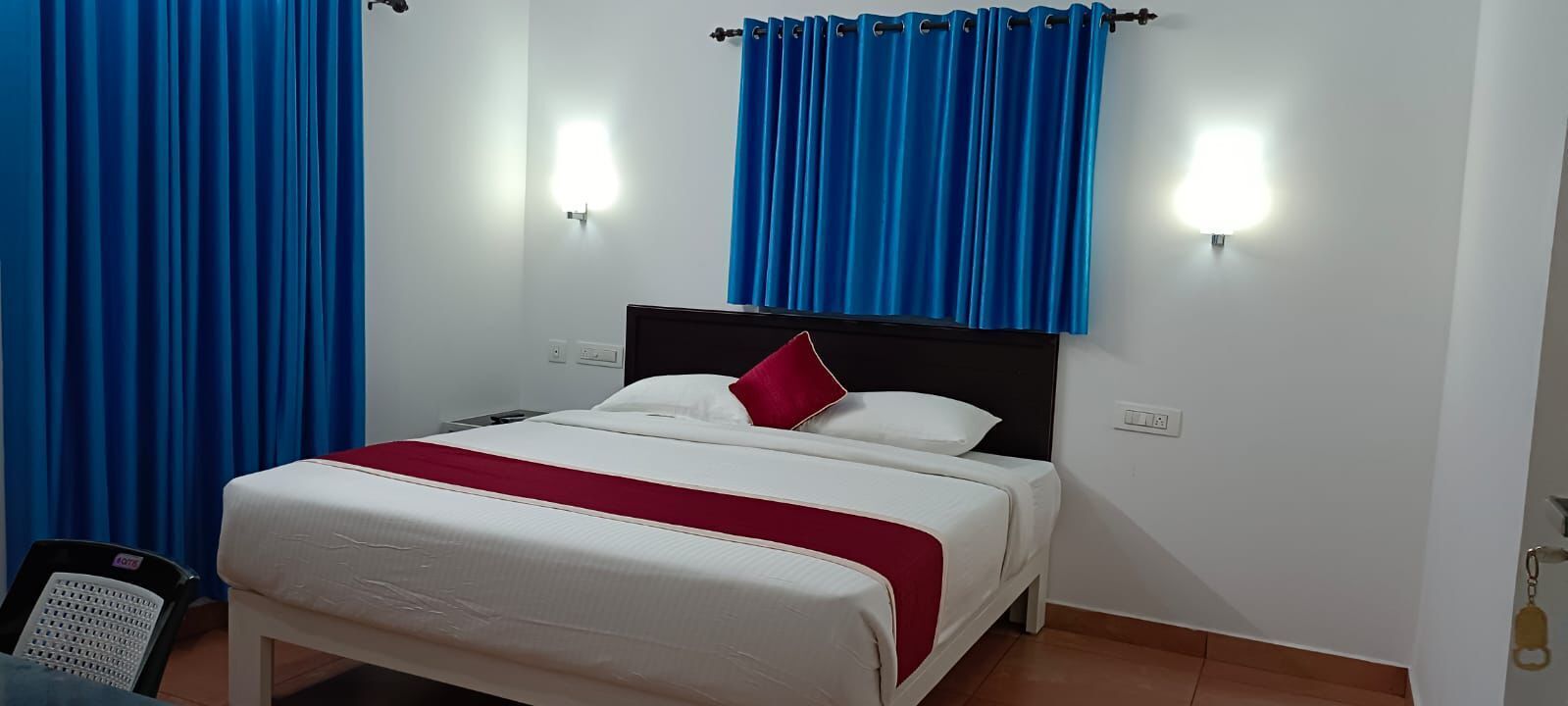 A neatly made bed with a red pillow, white bedding, and a black headboard. There are two wall-mounted lights on either side of the bed.