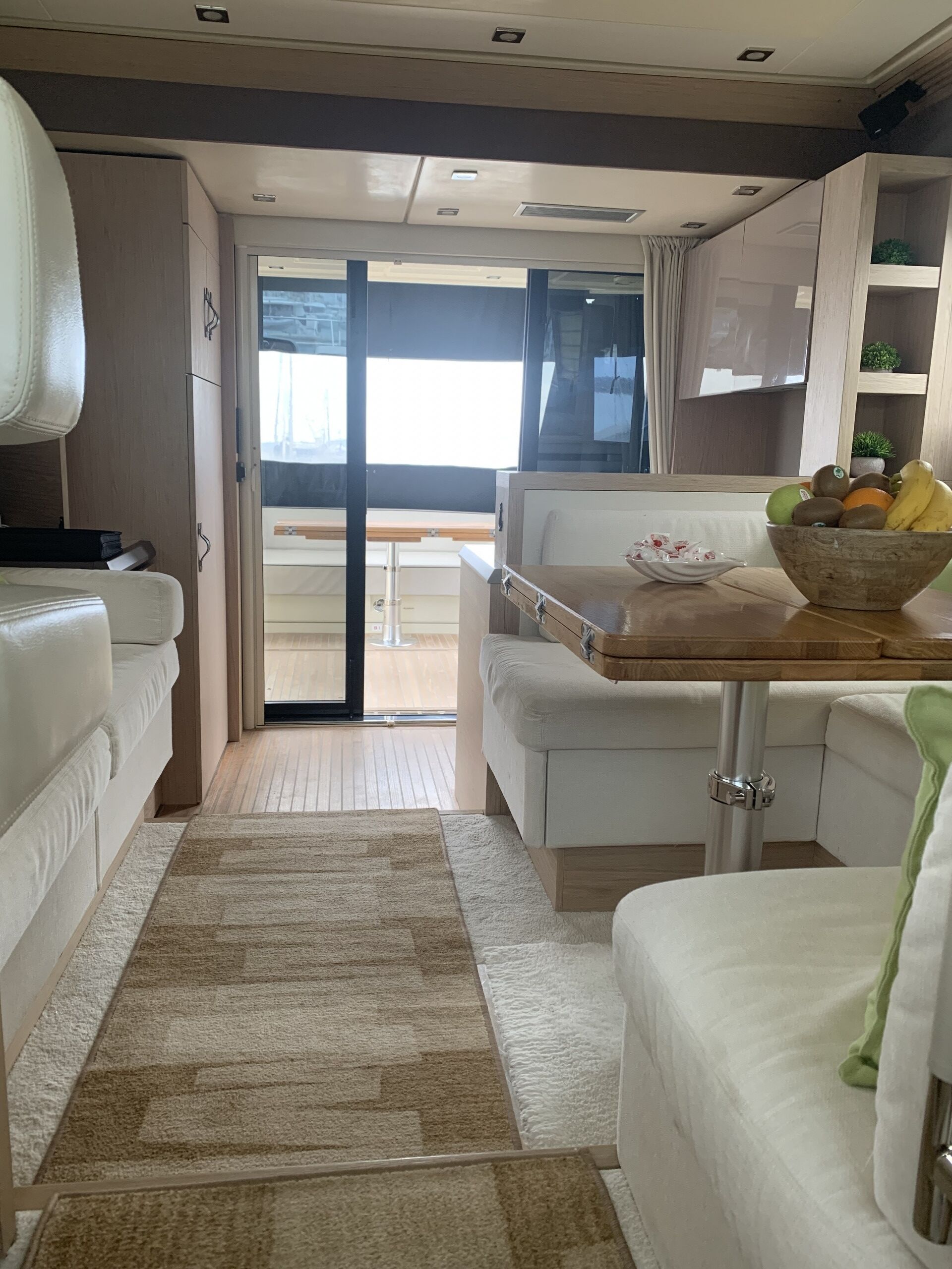 Een compact, goed verlicht interieur met een bank, een kleine eettafel met een schaal fruit en uitzicht door een glazen deur op een buitengebied.