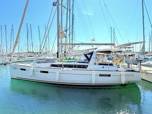 Eine weiße Yacht liegt in einer Marina festgemacht, wo sich weitere Boote befinden, der Himmel ist wolkenlos und es wehen Flaggen.