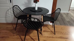 Une table de salle à manger ronde, accompagnée de deux chaises à armature métallique noire, un vase blanc contenant une plante et, au fond, une porte en bois.